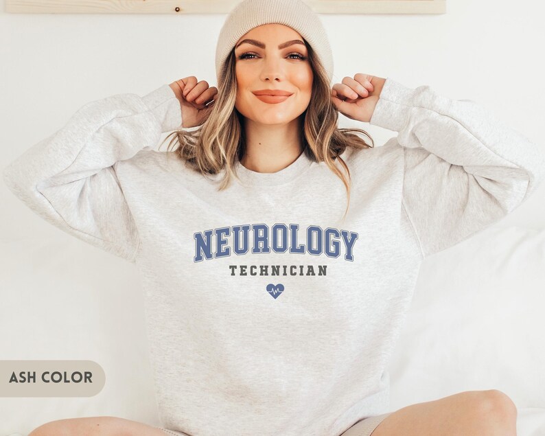 May include: A light gray sweatshirt with the text "Neurology Technician" in blue lettering and a small blue heart graphic. The sweatshirt is being worn by a person with long brown hair and a light gray beanie.