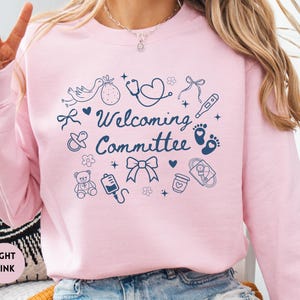 May include: Light pink sweatshirt with the words "Welcoming Committee" and various baby-themed illustrations in navy blue. The design includes a stork, baby feet, a stethoscope, and a teddy bear.