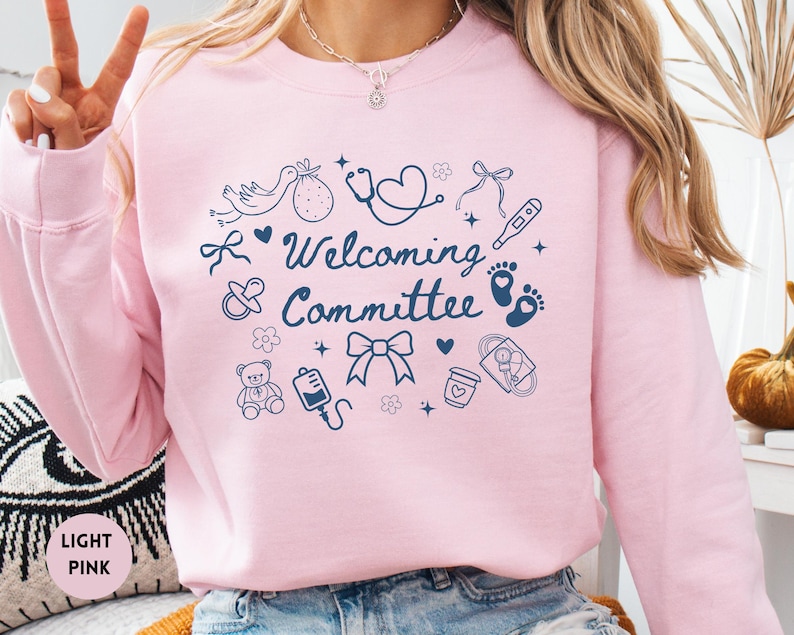 May include: Light pink sweatshirt with the words "Welcoming Committee" and various baby-themed illustrations in navy blue. The design includes a stork, baby feet, a stethoscope, and a teddy bear.