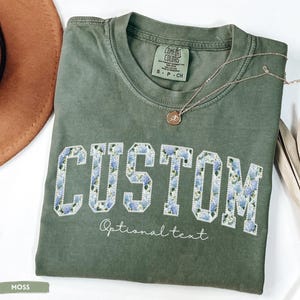 May include: A moss green t-shirt with the word "CUSTOM" in white block letters filled with a blue floral pattern. The text "Optional text" is below the word "CUSTOM". A gold necklace with a pendant is on the shirt.
