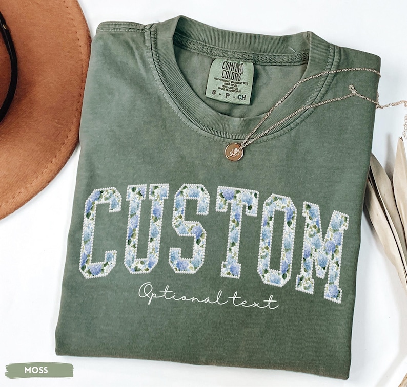 May include: A moss green t-shirt with the word "CUSTOM" in white block letters filled with a blue floral pattern. The text "Optional text" is below the word "CUSTOM". A gold necklace with a pendant is on the shirt.