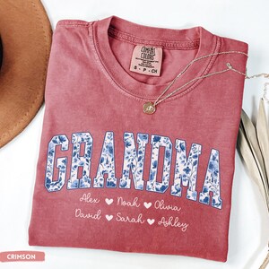 May include: Crimson t-shirt featuring the word "GRANDMA" in a blue floral design. Underneath are names: Alex, Noah, Olivia, David, Sarah, and Ashley. A gold pendant necklace rests on the shirt.
