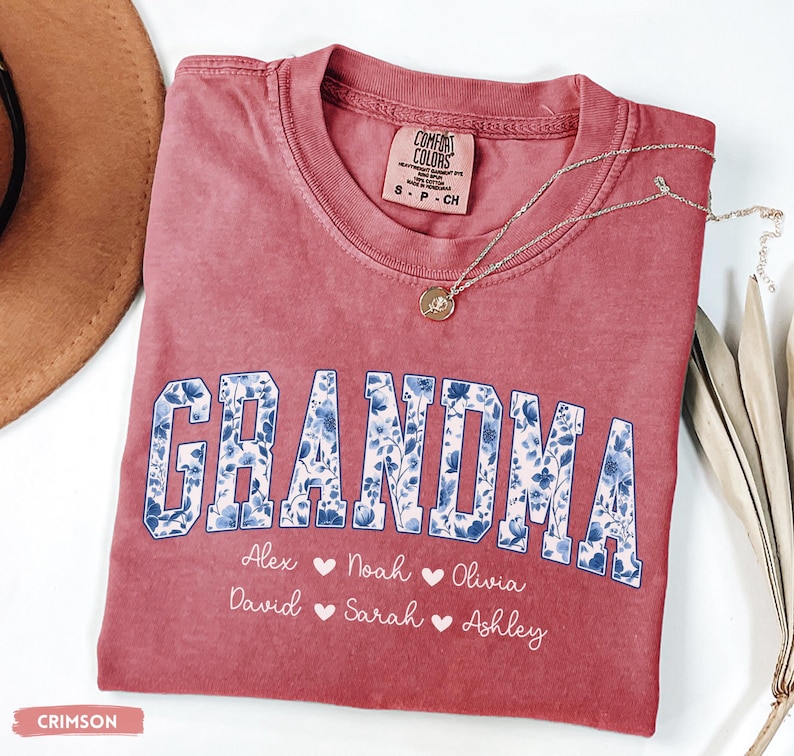 May include: Crimson t-shirt featuring the word "GRANDMA" in a blue floral design. Underneath are names: Alex, Noah, Olivia, David, Sarah, and Ashley. A gold pendant necklace rests on the shirt.
