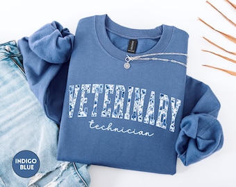 Puede incluir: Una sudadera azul de cuello redondo con la palabra VETERINARY en letras de bloque con un patrón floral azul y blanco, y la palabra technician en cursiva blanca debajo. Un collar de cadena plateada descansa sobre el cuello.