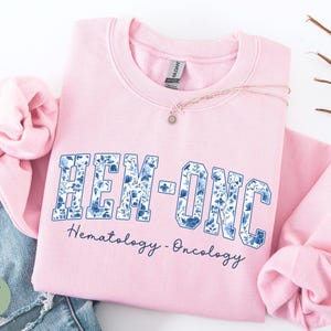 May include: A light pink sweatshirt with the text "HEM-ONC" in a blue floral design. Below the text is the phrase "Hematology - Oncology" in a dark blue script. A delicate necklace with a small pendant is visible.