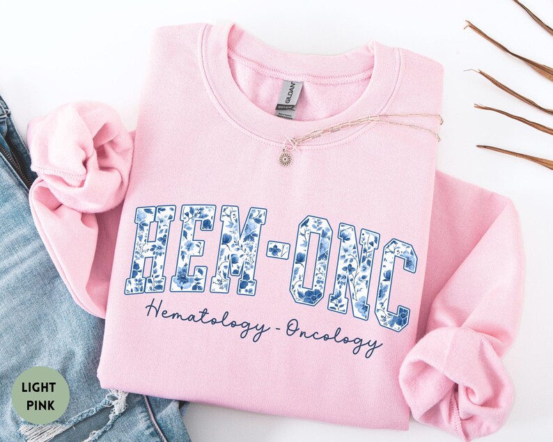 May include: A light pink sweatshirt with the text "HEM-ONC" in a blue floral design. Below the text is the phrase "Hematology - Oncology" in a dark blue script. A delicate necklace with a small pendant is visible.