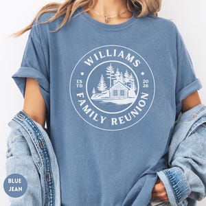 May include: A blue jean colored t-shirt featuring a white circular graphic on the chest. The design includes a cabin, pine trees, and the text WILLIAMS FAMILY REUNION ESTD 2026. The shirt has short sleeves and a crew neckline, worn with a denim jacket.