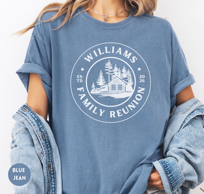 May include: A blue jean colored t-shirt featuring a white circular graphic on the chest. The design includes a cabin, pine trees, and the text WILLIAMS FAMILY REUNION ESTD 2026. The shirt has short sleeves and a crew neckline, worn with a denim jacket.