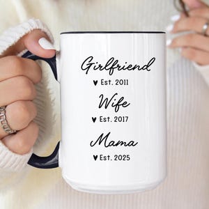 May include: White ceramic mug with black text that reads "Girlfriend Est. 2011, Wife Est. 2017, Mama Est. 2025".
