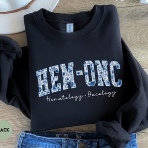 May include: Black sweatshirt with the text "HEM-ONC" in a blue floral design. The words "Hematology - Oncology" are written below in a script font. The sweatshirt is folded on top of blue jeans.