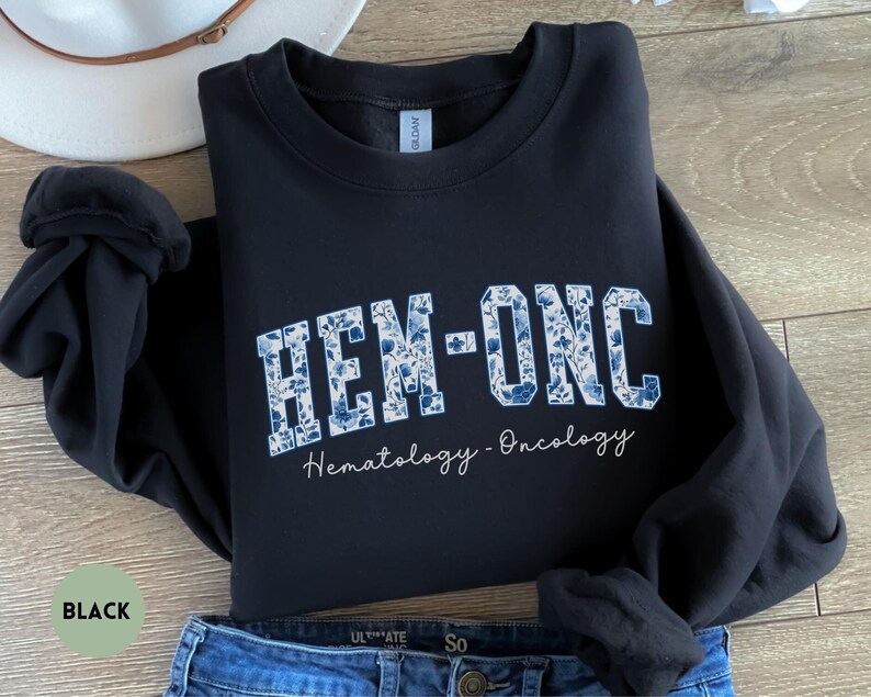 May include: Black sweatshirt with the text "HEM-ONC" in a blue floral design. The words "Hematology - Oncology" are written below in a script font. The sweatshirt is folded on top of blue jeans.
