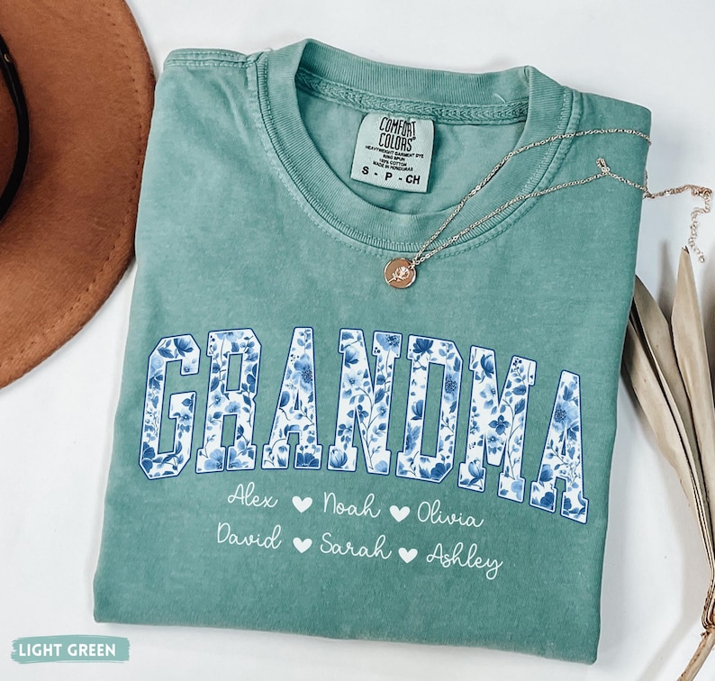 May include: A light green Comfort Colors t-shirt with the word "GRANDMA" in large, blue floral print letters. Below the word are names with heart symbols. A silver necklace with a pendant rests on the shirt. The shirt is folded.