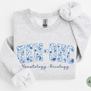 May include: Light gray sweatshirt with the text "HEM-ONC" in a blue floral design. Below the text is the phrase "Hematology - Oncology" in a script font. The sweatshirt has a crew neck and long sleeves.