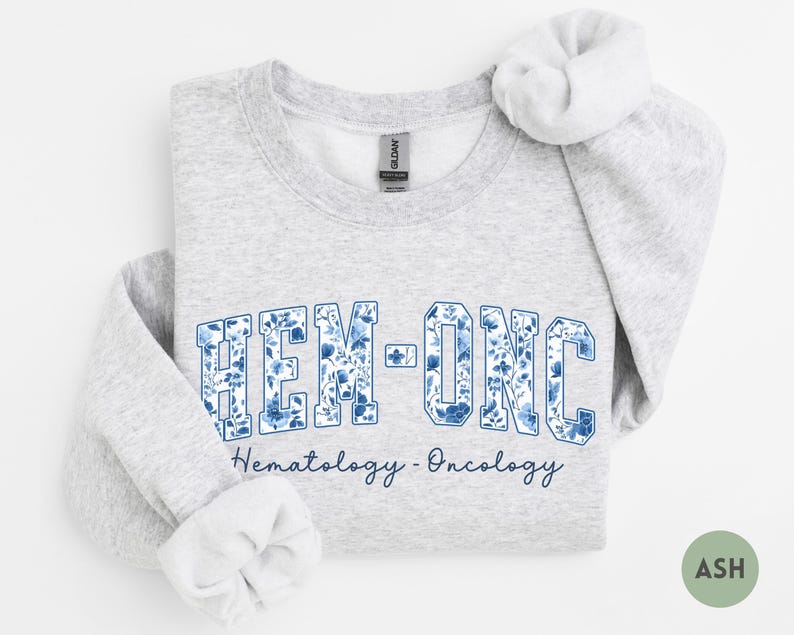 May include: Light gray sweatshirt with the text "HEM-ONC" in a blue floral design. Below the text is the phrase "Hematology - Oncology" in a script font. The sweatshirt has a crew neck and long sleeves.