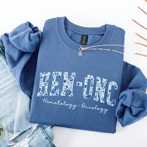 May include: Indigo blue sweatshirt with the text "HEM-ONC" in a floral design, with "Hematology-Oncology" written below. A silver necklace is draped across the sweatshirt, which is folded on top of a pair of blue jeans.