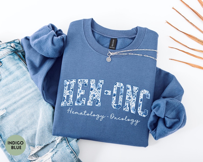 May include: Indigo blue sweatshirt with the text "HEM-ONC" in a floral design, with "Hematology-Oncology" written below. A silver necklace is draped across the sweatshirt, which is folded on top of a pair of blue jeans.