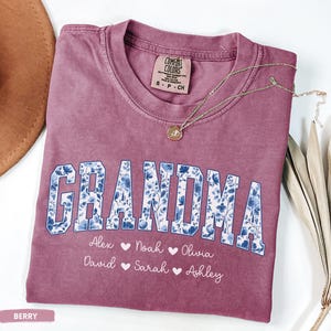 May include: Berry-coloured t-shirt with the word "GRANDMA" in a blue floral design. Below the word are the names Alex, Noah, Olivia, David, Sarah, and Ashley. A gold necklace with a pendant rests on the shirt.