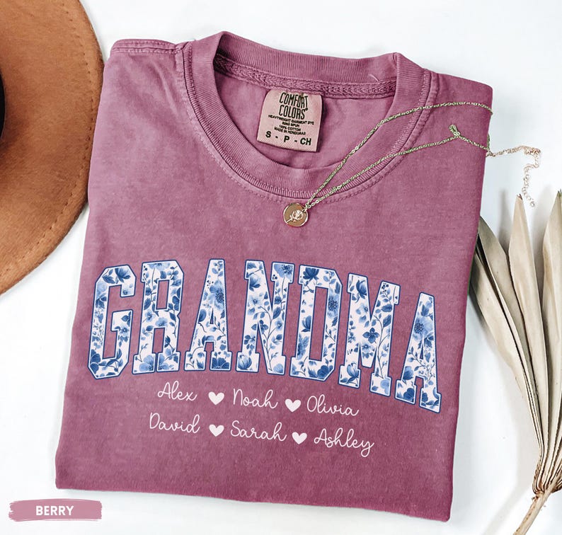 May include: Berry-coloured t-shirt with the word "GRANDMA" in a blue floral design. Below the word are the names Alex, Noah, Olivia, David, Sarah, and Ashley. A gold necklace with a pendant rests on the shirt.