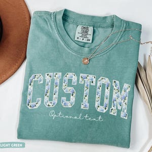 May include: A light green t-shirt with the word "CUSTOM" in a floral design. The shirt has the text "Optional text" below the word "CUSTOM". A silver necklace with a pendant is on the shirt.