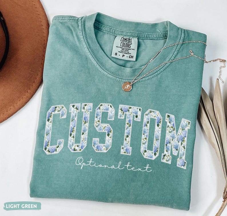 May include: A light green t-shirt with the word "CUSTOM" in a floral design. The shirt has the text "Optional text" below the word "CUSTOM". A silver necklace with a pendant is on the shirt.