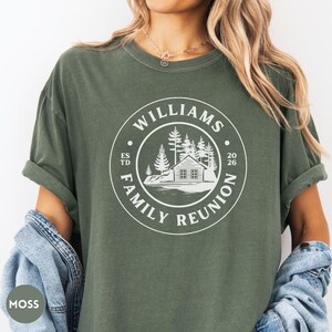 May include: A moss green short-sleeve t-shirt featuring a white circular graphic on the chest. The design includes a cabin, pine trees, and the text "WILLIAMS FAMILY REUNION" along with "ESTD 2026" printed in a clean, sans-serif font style.
