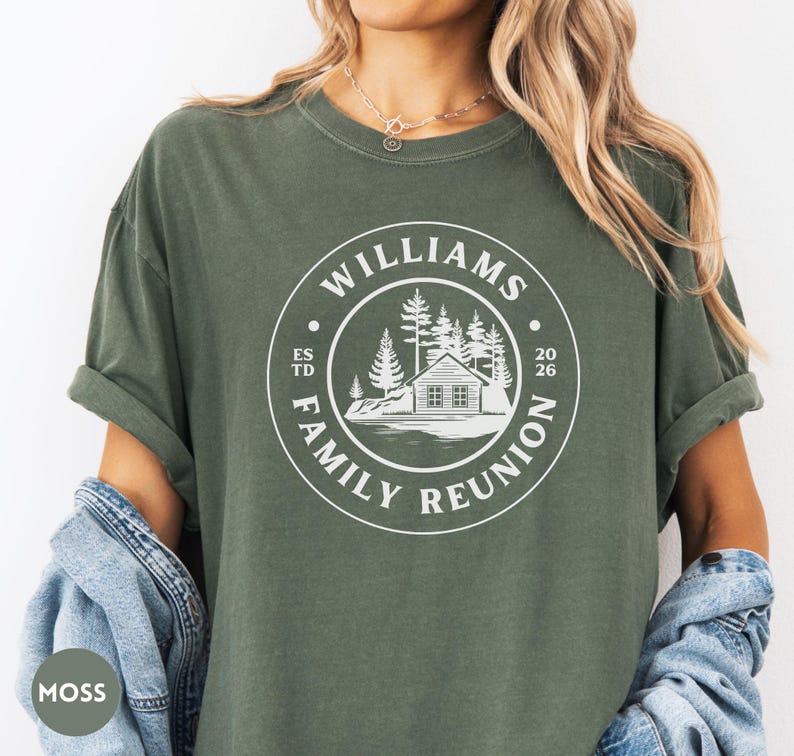 May include: A moss green short-sleeve t-shirt featuring a white circular graphic on the chest. The design includes a cabin, pine trees, and the text "WILLIAMS FAMILY REUNION" along with "ESTD 2026" printed in a clean, sans-serif font style.