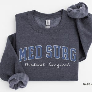 May include: Dark heather gray sweatshirt with the text "MED SURG" in blue block letters with a white outline. Below the text is the phrase "Medical - Surgical" in a cursive font. The sweatshirt is folded.