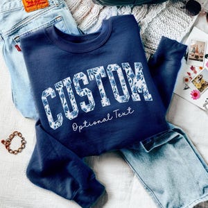 May include: A navy blue long-sleeve sweatshirt featuring the word CUSTOM in large block letters filled with a blue and white floral pattern. Below the word, the text Optional Text is written in a white cursive script font on the chest area.
