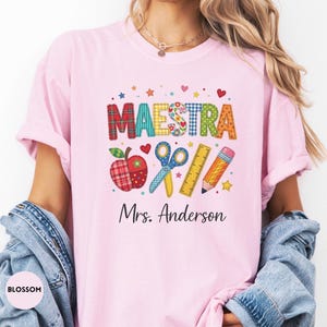 May include: A light pink t-shirt featuring a graphic print with the word MAESTRA in colorful patterned letters, an apple, scissors, a ruler, and a pencil. Below the graphic, the text Mrs. Anderson is printed in a black cursive script font.