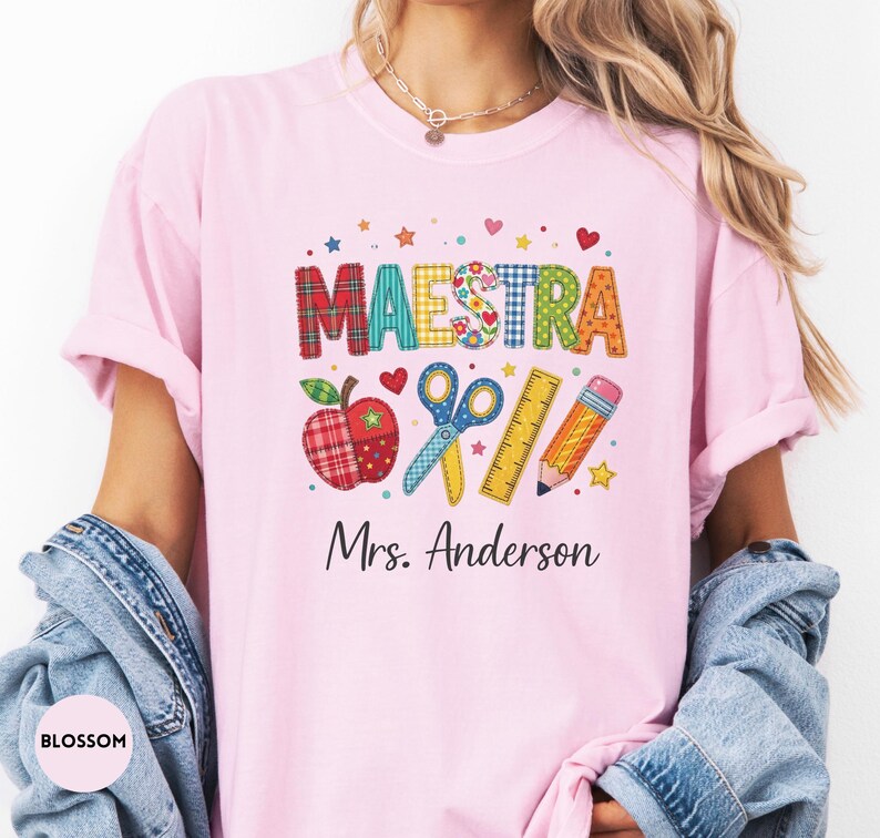 May include: A light pink t-shirt featuring a graphic print with the word MAESTRA in colorful patterned letters, an apple, scissors, a ruler, and a pencil. Below the graphic, the text Mrs. Anderson is printed in a black cursive script font.
