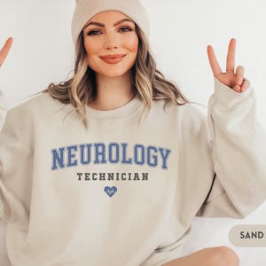 May include: A sand-colored sweatshirt with the text "NEUROLOGY TECHNICIAN" in blue lettering and a small blue heart with a heartbeat symbol.