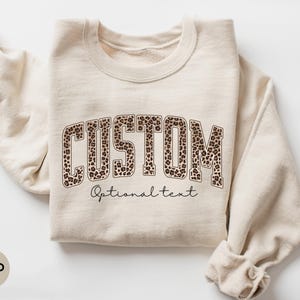 May include: Sand-colored sweatshirt with the word "CUSTOM" in a leopard print design. Below the word "CUSTOM" is the text "Optional text" in a script font. The sweatshirt is folded, with the sleeves visible.