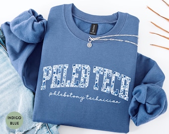 May include: Indigo blue sweatshirt with the text "PHLEB TECH" in white block letters filled with a blue floral pattern. The words "phlebotomy technician" are written below in a script font. A silver necklace is draped across the top.
