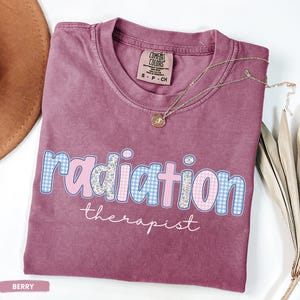 May include: Berry-coloured t-shirt featuring the words "radiation therapist". The word "radiation" is in a blue and pink gingham pattern with floral accents. A gold necklace with a pendant rests on the shirt.