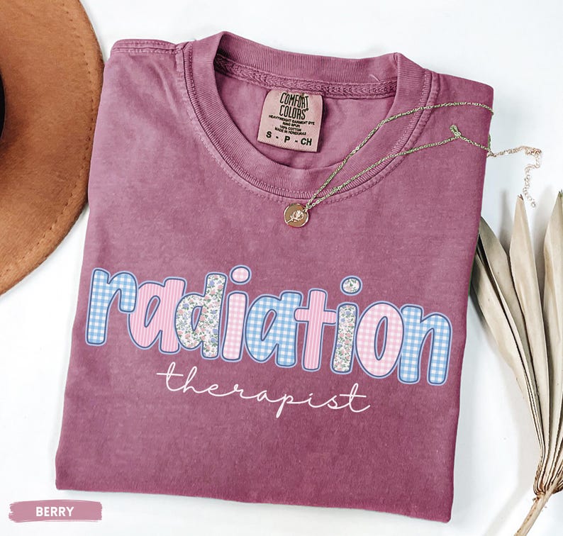May include: Berry-coloured t-shirt featuring the words "radiation therapist". The word "radiation" is in a blue and pink gingham pattern with floral accents. A gold necklace with a pendant rests on the shirt.
