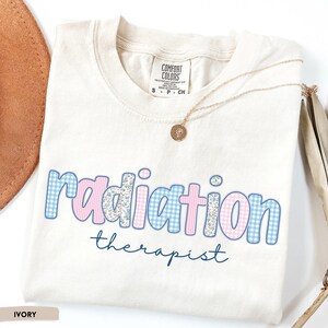 May include: Cream-coloured t-shirt featuring the words "radiation therapist". The word "radiation" is in pink and blue gingham and floral patterns. A gold necklace with a pendant rests on the shirt.