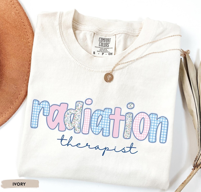 May include: Cream-coloured t-shirt featuring the words "radiation therapist". The word "radiation" is in pink and blue gingham and floral patterns. A gold necklace with a pendant rests on the shirt.