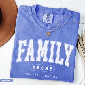 Peut inclure: Un t-shirt &agrave; manches courtes bleu pervenche avec une encolure ronde. Le devant pr&eacute;sente le texte blanc FAMILY dans une grande police universitaire, suivi de VACAY dans un contour ovale, et les mots CUSTOM LOCATION imprim&eacute;s en blanc.