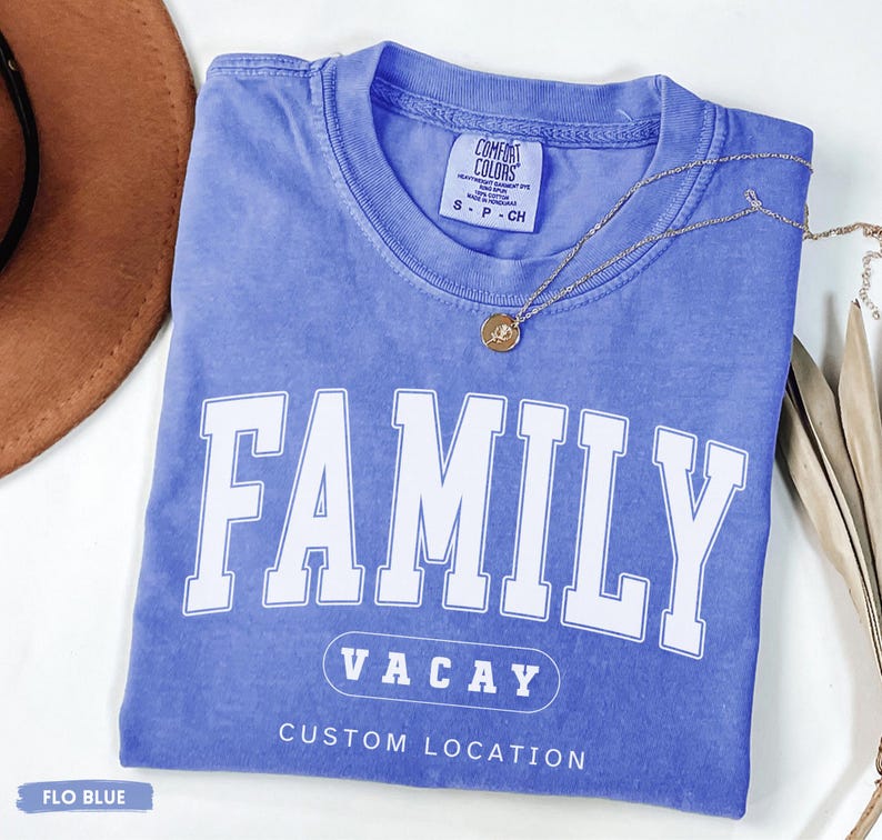 Peut inclure: Un t-shirt &agrave; manches courtes bleu pervenche avec une encolure ronde. Le devant pr&eacute;sente le texte blanc FAMILY dans une grande police universitaire, suivi de VACAY dans un contour ovale, et les mots CUSTOM LOCATION imprim&eacute;s en blanc.