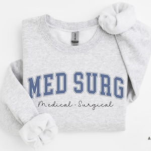 May include: A light gray sweatshirt with the words "MED SURG" in blue block letters. Below the text is the phrase "Medical - Surgical" in a script font. The sweatshirt is folded with the sleeves rolled up.
