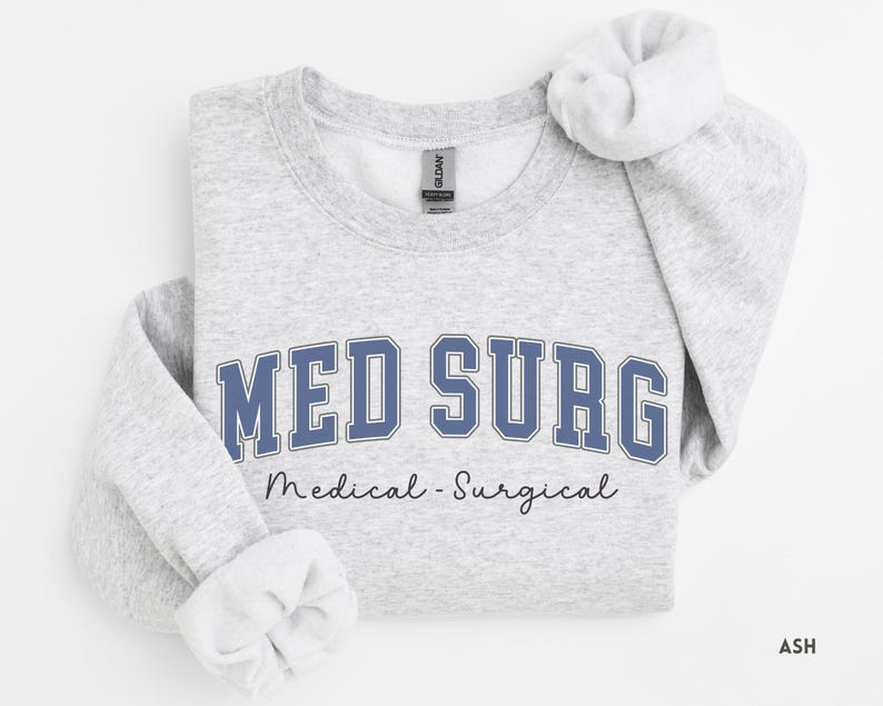 May include: A light gray sweatshirt with the words "MED SURG" in blue block letters. Below the text is the phrase "Medical - Surgical" in a script font. The sweatshirt is folded with the sleeves rolled up.