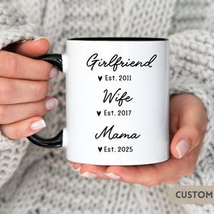 May include: White ceramic mug with a black handle and black rim. The mug has black text that reads "Girlfriend Est. 2011 Wife Est. 2017 Mama Est. 2025".