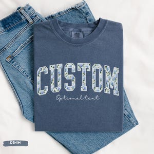 May include: A denim blue t-shirt with the word "CUSTOM" in a floral design. The text "Optional text" is below the word "CUSTOM". The t-shirt is folded on top of a pair of blue jeans.