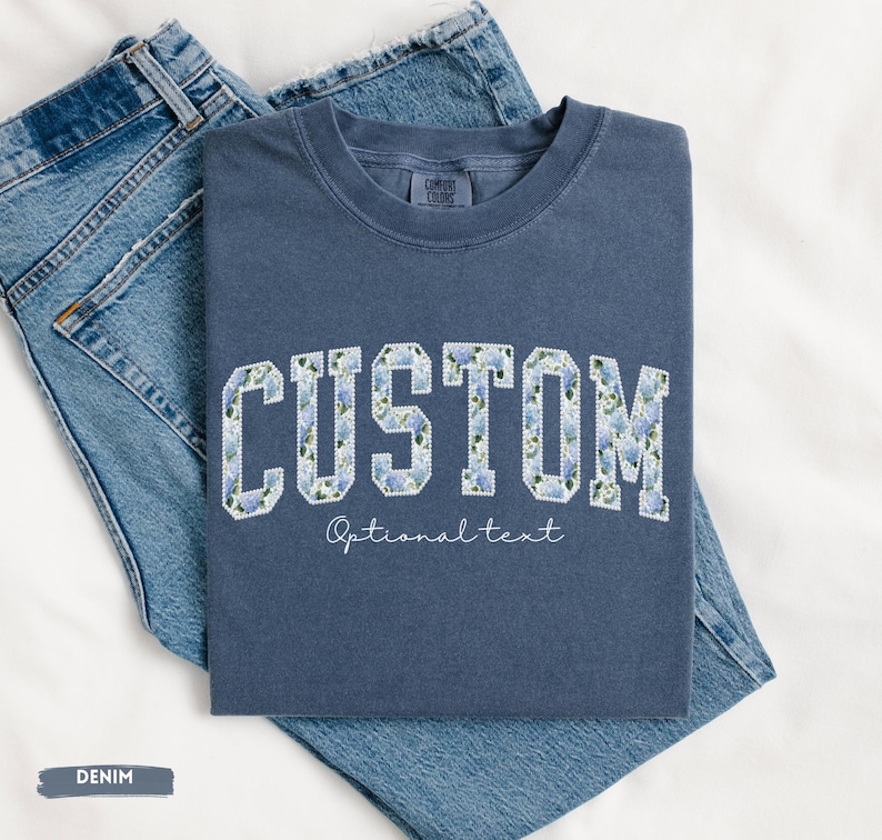 May include: A denim blue t-shirt with the word "CUSTOM" in a floral design. The text "Optional text" is below the word "CUSTOM". The t-shirt is folded on top of a pair of blue jeans.