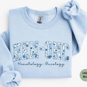 May include: A light blue sweatshirt with the text "HEM-ONC" in a floral design. Below the text is the phrase "Hematology - Oncology" in a script font. The sweatshirt has a crew neck and long sleeves.