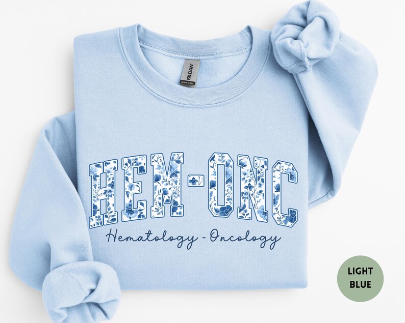 May include: A light blue sweatshirt with the text "HEM-ONC" in a floral design. Below the text is the phrase "Hematology - Oncology" in a script font. The sweatshirt has a crew neck and long sleeves.