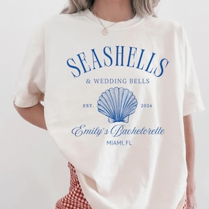 May include: Cream-colored t-shirt with blue text that reads "Seashells & Wedding Bells" above a seashell graphic. Below the graphic, it says "Emily's Bachelorette, Miami, FL" and "Est. 2026".