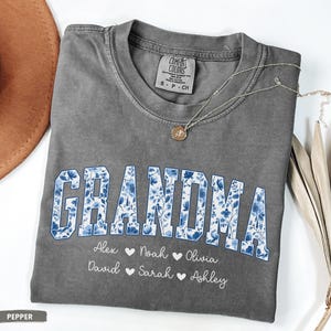 May include: A grey Comfort Colors t-shirt with the word "GRANDMA" in blue floral print. Below the word are names: Alex, Noah, Olivia, David, Sarah, and Ashley. A gold necklace with a pendant is on the shirt.