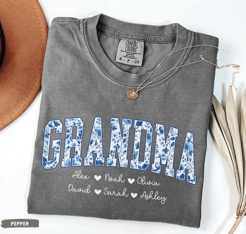 May include: A grey Comfort Colors t-shirt with the word "GRANDMA" in blue floral print. Below the word are names: Alex, Noah, Olivia, David, Sarah, and Ashley. A gold necklace with a pendant is on the shirt.