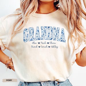 May include: Cream-coloured sweatshirt featuring the word "GRANDMA" in a blue floral design. Underneath are the names Alex, Noah, Olivia, David, Sarah, and Ashley, each with a heart symbol. The word "IVORY" is printed at the bottom.